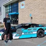 Race car driver wearing all black stands in front of blue, black and silver race car parked in front of large brick building with Kids' Food Basket logo.