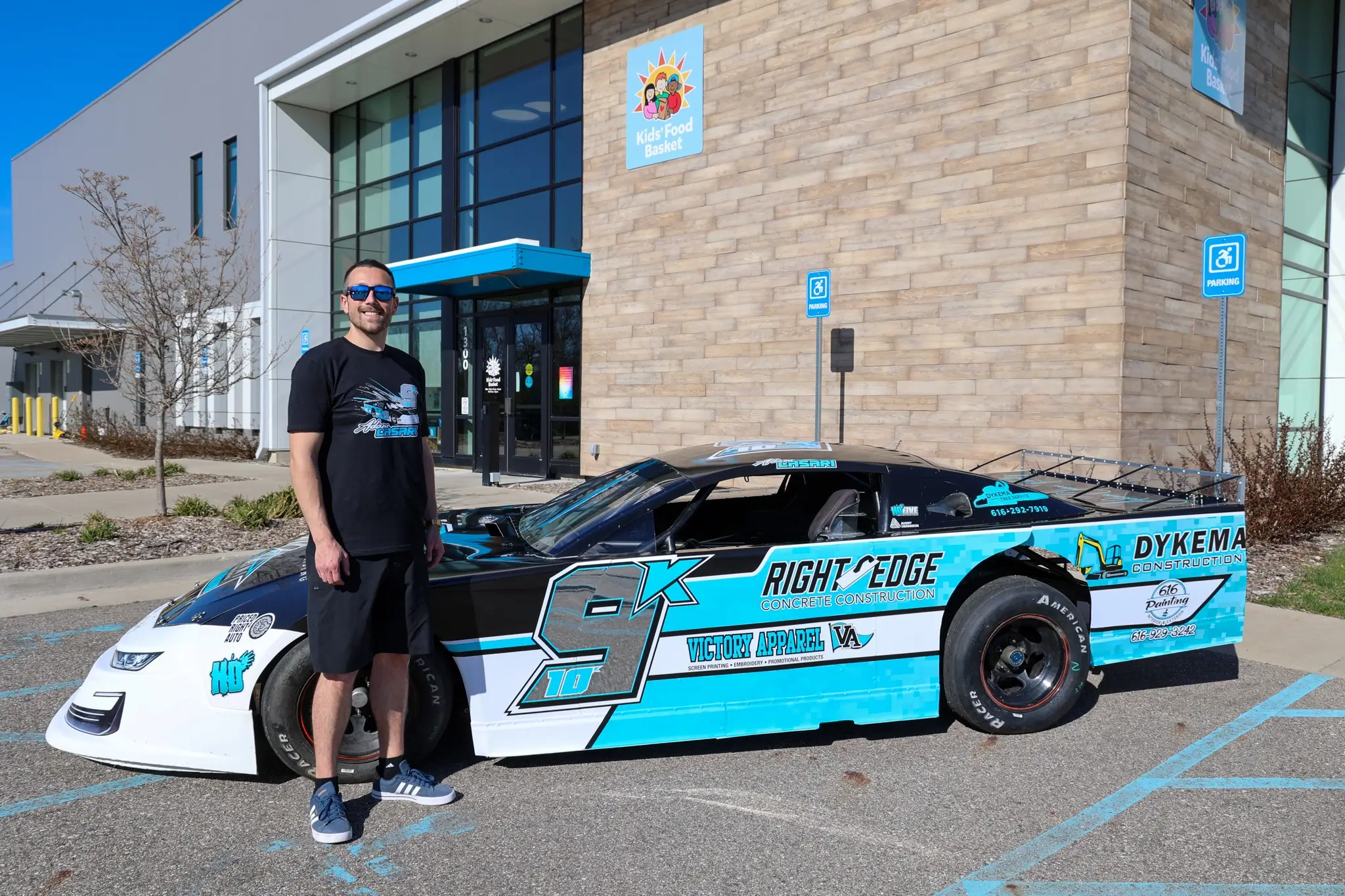 Race car driver wearing all black stands in front of blue, black and silver race car parked in front of large brick building with Kids' Food Basket logo.