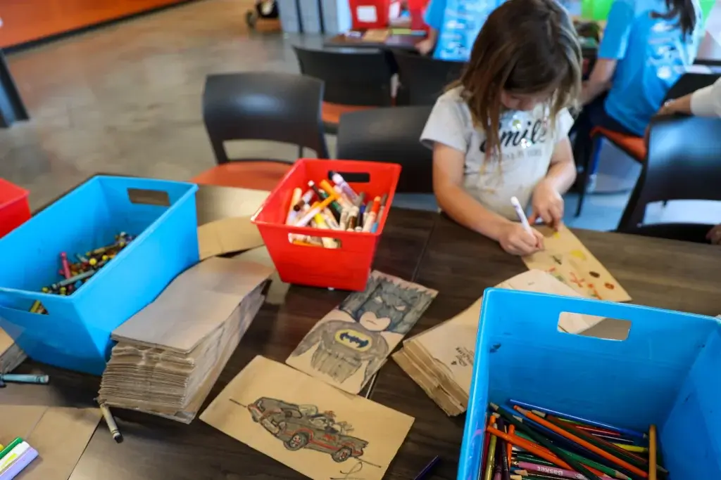 A child at a brown table bent over a brown paper bag, decorating with marker.