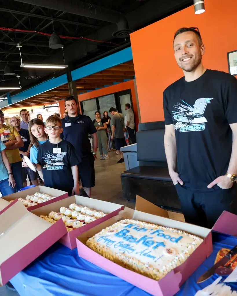 Race car driver Adam Casari stands in front of a birthday cake and cupcakes with friends, family and fans in the background.