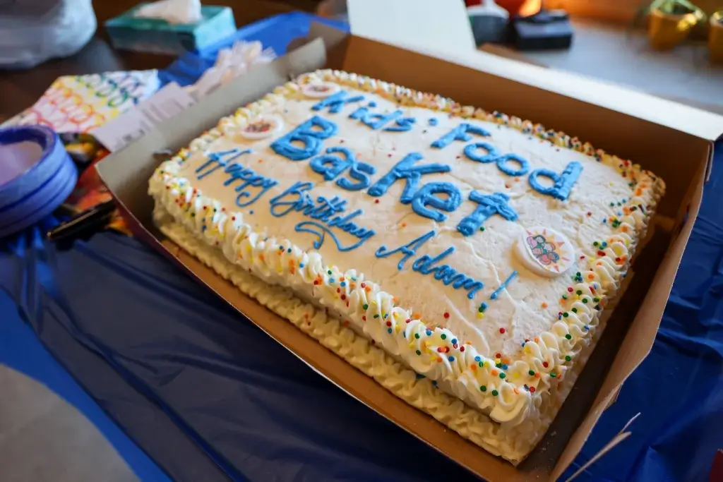 A box is open to reveal a white frosted cake with "Kids' Food Basket Happy Birthday Adam" written in blue frosting.