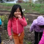 A child with long dark hair wearing a pink zippered coat smiles while nibbling a yellow flower. The child is surrounded by four other children exploring the grounds.