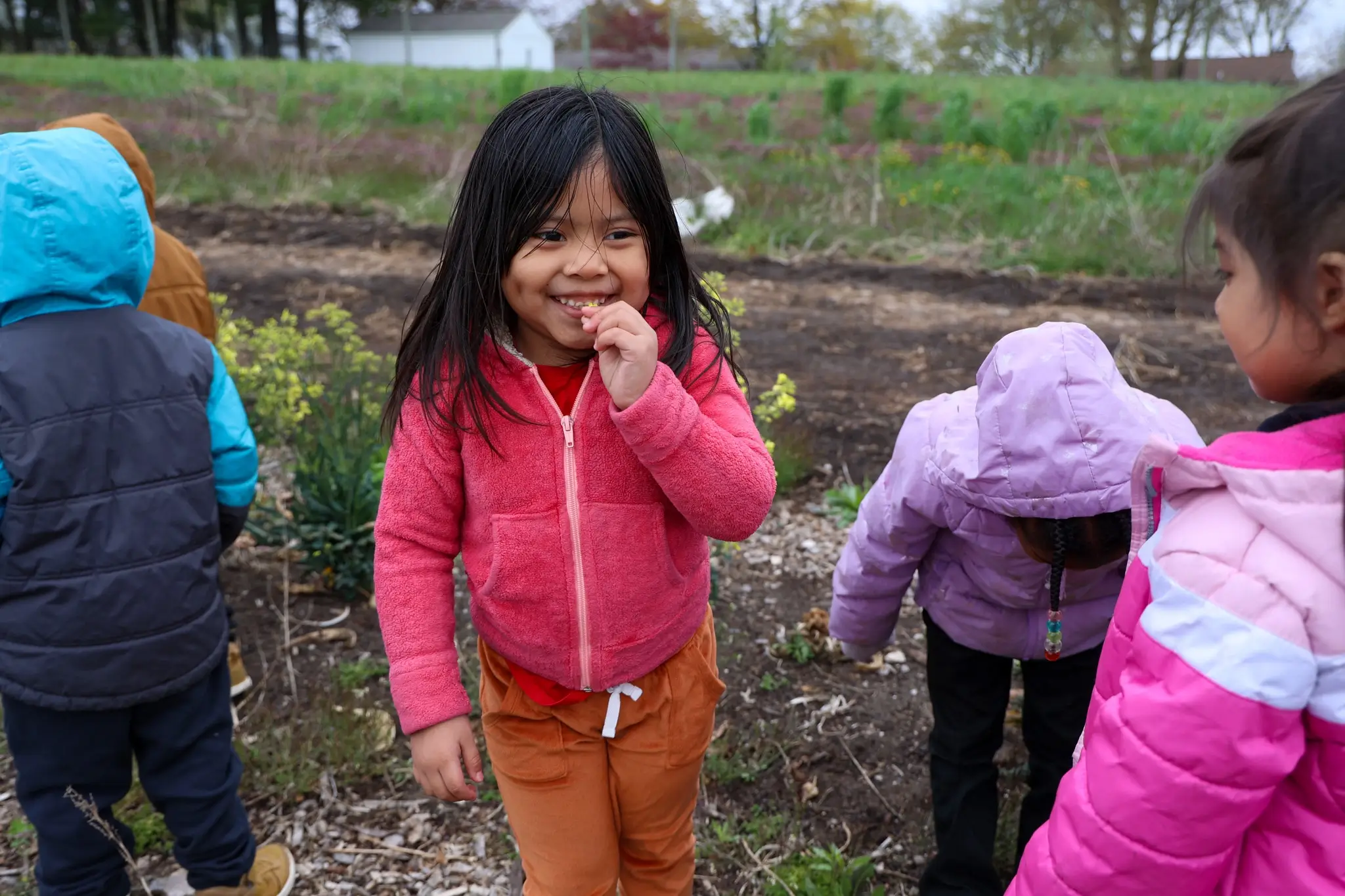 A child with long dark hair wearing a pink zippered coat smiles while nibbling a yellow flower. The child is surrounded by four other children exploring the grounds.